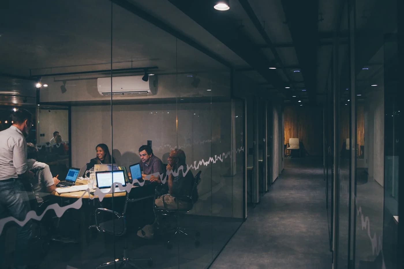 A team meeting in a glass-walled room, laptops open on the table, in an office at night.