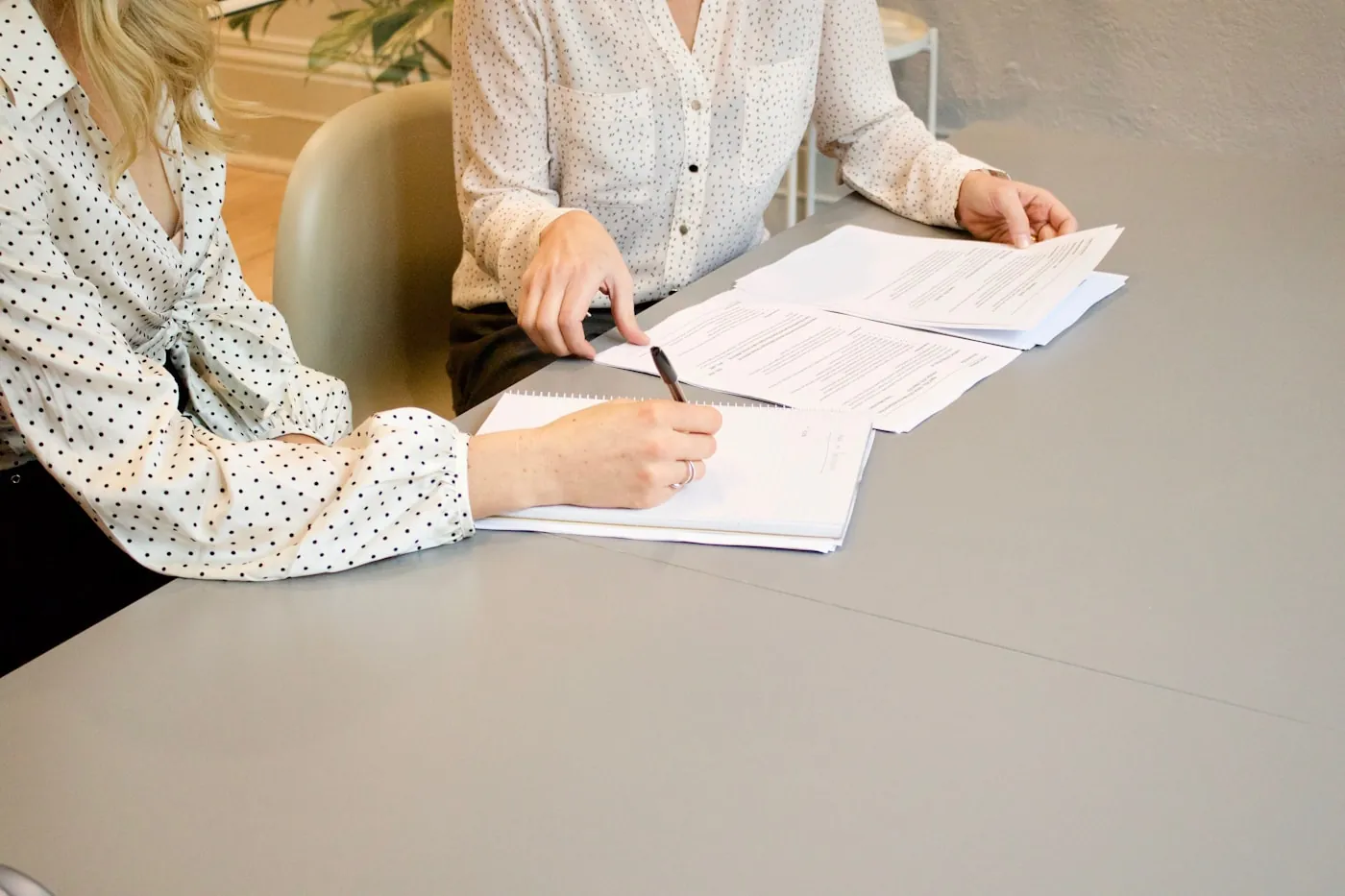 Two people at an office table with a printed contract open between them, one of them taking notes in a notebook.